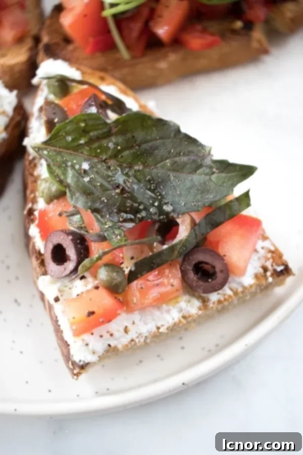 A gourmet bruschetta piece, elegantly topped with creamy ricotta, savory black olives, fresh diced tomatoes, and distinctive red basil leaves on toasted bread.
