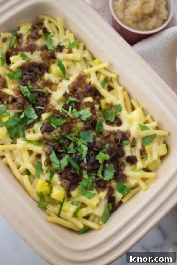 A delicious close-up of Älplermagronen, showcasing layers of golden macaroni, melted cheese, and soft potatoes, topped with caramelized onions and fresh parsley, with a small bowl of applesauce beside it.