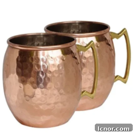 Two shiny copper Moscow Mule mugs, reflecting light, with limes suggesting the drink ingredients.